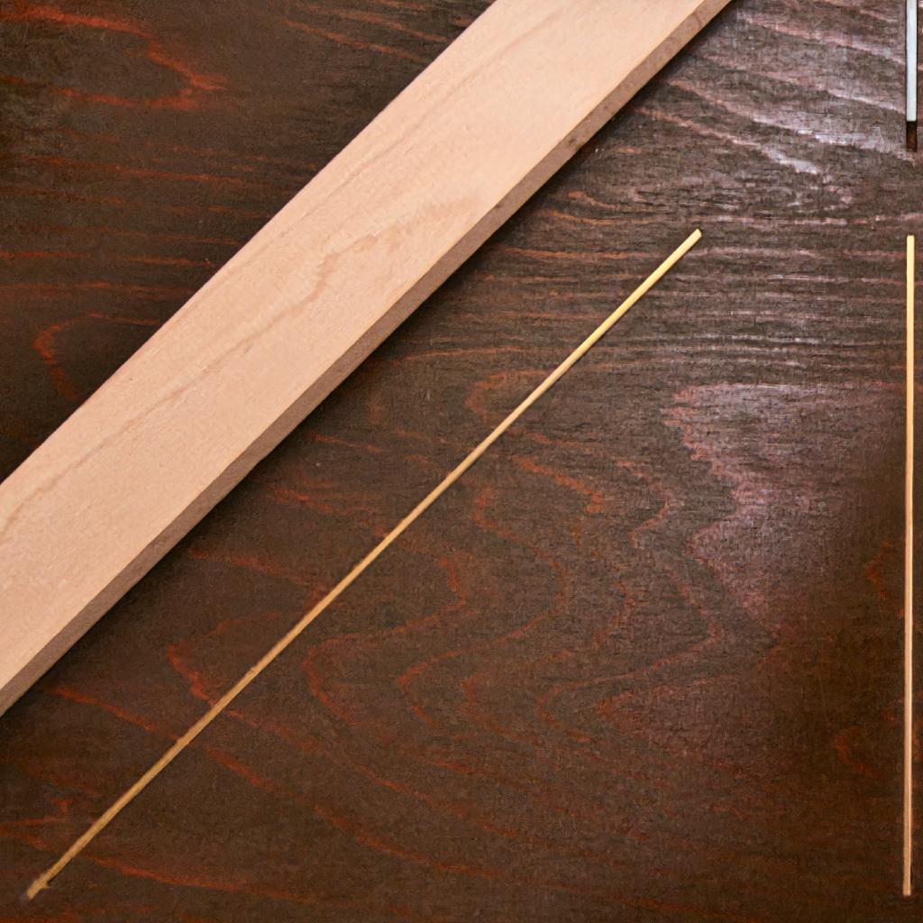Die abstrakte Nahaufnahme zeigt eine leicht rauhe Holzoberfläche aus dunkel lasiertem Holz mit einem langen unbehandelten Balken aus glattem, hellerem Holz, der auf dem dunklen Holz aufliegt und diagonal und nach oben versetzt von unten links nach oben rechts aus dem Bild läuft sowie zwei dünnen, goldfarbenen Stäben, die in das dunkle Holz eingelassen sind. Einer verläuft parallel unter dem Holzbalken und beginnt knapp über der unteren, linken Ecke, stoppt aber deutlich vor der oberen rechten Ecke. Der zweite verläuft parallel zur rechten Kante des Fotos, wobei dieser oben rechts kurz unterbrochen ist. Das Foto ist im 1:1-Format geschnitten.