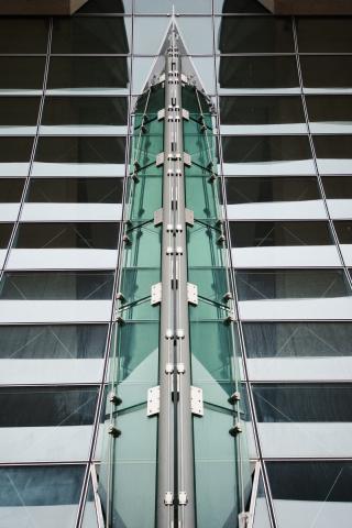 Das Foto zeigt eine moderne Architekturaufnahme, die sich durch symmetrische Linien und die Verwendung von Glas und Metall auszeichnet. Die Komposition ist streng symmetrisch aufgebaut und zeigt die Fassade eines Gebäudes. In der Mitte verläuft eine vertikale Struktur, die von zwei Fassadenteilen flankiert wird. Die Fassadenteile sind durch horizontale Linien unterteilt, die Fensterreihen bilden. Die vertikale Struktur scheint aus Glas und Metall zu bestehen und bildet den Schwerpunkt des Bildes, wobei die horizontale und diagonale Linien der Fassade ein starkes visuelles Muster erzeugen, das den Blick des Betrachters lenkt. Die Aufnahme ist frontal, was die Symmetrie und Geometrie der Architektur zusätzlich betont.