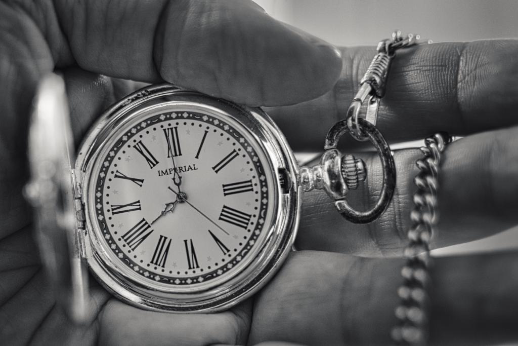 Das Schwarz-Weiß-Bild zeigt eine Nahaufnahme einer Hand, die eine silberne Taschenuhr mit geöffnetem Deckel und Kette hält, wobei der Fokus auf dem detailreichen Zifferblatt liegt. Die Komposition ist nah und fokussiert, wobei die Uhr zentral im Bild gehalten wird. Der Betrachter sieht die Uhr mit geöffnetem Deckel, was ihr Zifferblatt vollständig sichtbar macht. Die Hand, die die Uhr hält, ist leicht unscharf, was den Fokus auf die Uhr selbst lenkt. Die Kette der Uhr liegt locker neben dem Uhrwerk, was einen zusätzlichen dekorativen Akzent liefert. Das Bild scheint im Moment zu verharren, wobei die Zeit auf dem Zifferblatt stillsteht. Das zentrale Element ist die Taschenuhr, die durch ihr silbernes Metall und das klare weiße Zifferblatt mit schwarzen römischen Ziffern und filigranen Zeigern auffällt. Auf dem Zifferblatt steht die Marke "IMPERIAL". Die Hand, die die Uhr hält, ist von mittlerem Teint, die Details der Hautstruktur sind erkennbar. Der Daumen ist leicht angewinkelt.