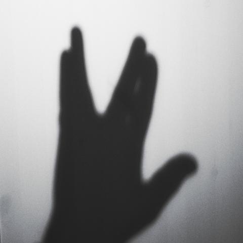 Die Schwarz-Weiß-Fotografie zeigt den Schatten einer Hand, die das vulkanische Grußzeichen aus der Fernsehserie Star Trek formt, vor einem weißen Hintergrund. Die Komposition ist einfach und minimalistisch, wobei der Fokus ausschließlich auf dem Schatten der Hand liegt. Die Pose der Hand, mit gespreizten Fingern, ist deutlich erkennbar, und der Betrachter ist sofort in der Lage, das berühmte Star Trek-Zeichen zu erkennen. Der Hintergrund ist einfarbig weiß, wodurch der Schatten der Hand klar und deutlich hervorgehoben wird.