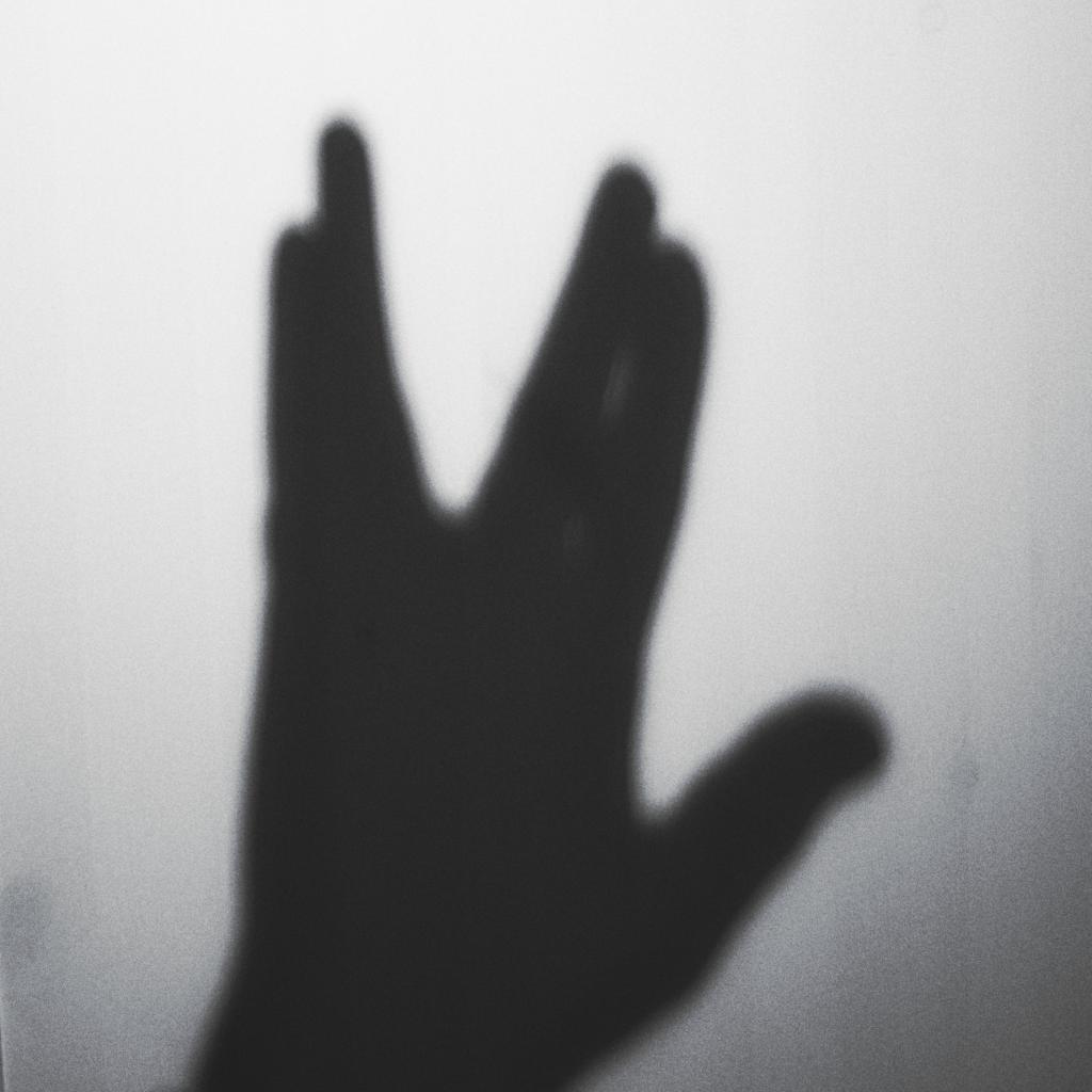 Die Schwarz-Weiß-Fotografie zeigt den Schatten einer Hand, die das vulkanische Grußzeichen aus der Fernsehserie Star Trek formt, vor einem weißen Hintergrund. Die Komposition ist einfach und minimalistisch, wobei der Fokus ausschließlich auf dem Schatten der Hand liegt. Die Pose der Hand, mit gespreizten Fingern, ist deutlich erkennbar, und der Betrachter ist sofort in der Lage, das berühmte Star Trek-Zeichen zu erkennen. Der Hintergrund ist einfarbig weiß, wodurch der Schatten der Hand klar und deutlich hervorgehoben wird.