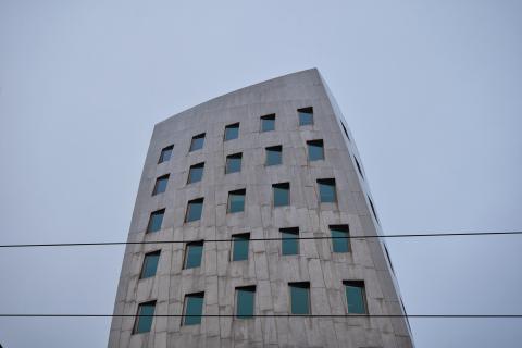 Das Bild zeigt ein modernes, hoch aufragendes Gebäude aus grauem, glattem Stahlbeton unter einem graublauen Himmel. Die Komposition des Fotos ist durch eine leicht untersichtige Perspektive gekennzeichnet, wobei das Gebäude im Mittelpunkt steht und sich leicht nach oben neigt, was seine Höhe betont. Die Fenster sind nicht horizontal angeordnet, sondern leicht nach rechts ansteigend. Im Vordergrund verlaufen Stromleitungen, die einen zusätzlichen Kontrast zum strengen geometrischen Design des Gebäudes bilden.