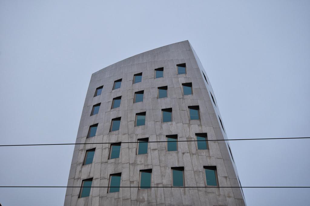 Das Bild zeigt ein modernes, hoch aufragendes Gebäude aus grauem, glattem Stahlbeton unter einem graublauen Himmel. Die Komposition des Fotos ist durch eine leicht untersichtige Perspektive gekennzeichnet, wobei das Gebäude im Mittelpunkt steht und sich leicht nach oben neigt, was seine Höhe betont. Die Fenster sind nicht horizontal angeordnet, sondern leicht nach rechts ansteigend. Im Vordergrund verlaufen Stromleitungen, die einen zusätzlichen Kontrast zum strengen geometrischen Design des Gebäudes bilden.