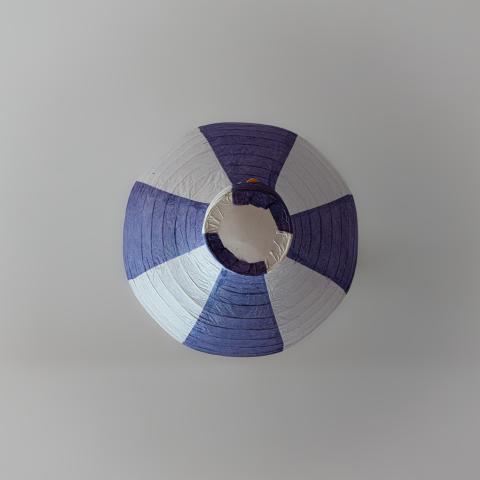 Das Bild zeigt eine Papierlampe in einem Froschperspektiven-Blick, mit abwechselnd blauvioletten und weißen Segmenten, die von der weißen Decke hängt. Die Komposition präsentiert die Lampe zentral, von oben fotografiert, wobei die Kamera direkt auf die Unterseite der Lampe gerichtet ist. Die blauen und weißen Segmente erstrecken sich vom Zentrum nach außen und bilden ein kreisförmiges Muster. Der Hintergrund besteht aus einem gleichmäßigen, neutralen Grau-Weiß, das die Laterne hervorhebt. Die Beleuchtung ist gleichmäßig, ohne deutliche Schatten, wodurch ein minimalistischer und fast abstrakter Look entsteht.