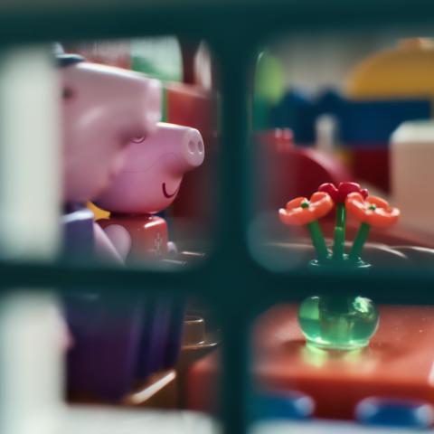 Das Bild fängt eine intime Nahaufnahme von Spielzeugfiguren im Fokus ein, wobei der Betrachter durch ein Fenster oder eine ähnliche Struktur blickt. Die Szene wird durch die Rahmung der Aufnahme strukturiert, wobei eine grüne, gitterartige Struktur im Vordergrund als Fenster oder Rahmen dient, durch das der Betrachter auf die eigentliche Szene blickt. Die Hauptfiguren, zwei Spielzeugfiguren, die an Peppa-Pig-Figuren im Lego-Duplo-Look erinnern, befinden sich im Hintergrund, umgeben von bunten Legosteinen. Im rechten Bereich des Bildes stehen kleine, rote Blüten in einer grünen Vase, die auf einer roten Oberfläche positioniert sind. Die Komposition erzeugt eine visuelle Trennung, die den Betrachter durch das Fenster in das Spielzeug-Universum einlädt. Die unscharfe Darstellung des Hintergrundes lenkt die Aufmerksamkeit auf die Hauptfiguren und die Blumen.