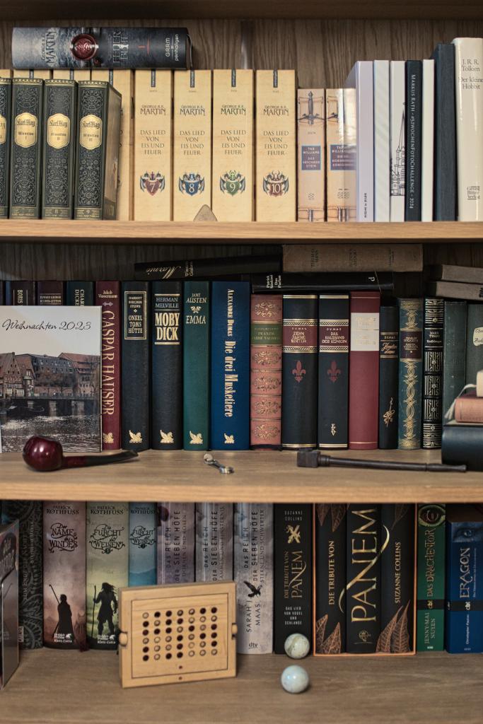 Ein Bücherregal mit mehreren Regalen voller Bücher in verschiedenen Größen, Farben und Einbänden. Auf dem obersten Regal stehen vor allem ältere, gebundene Bücher mit goldenen Verzierungen auf den Buchrücken. Darunter befindet sich eine Reihe von Büchern mit dem Titel "Das Lied von Eis und Feuer", erkennbar an ihren farbigen Buchrücken und stilisierten Emblemen. Das mittlere Regal zeigt eine buntere Mischung, darunter "Moby Dick", "Die drei Musketiere" und einige Bücher mit roten, grünen und braunen Einbänden. Links auf diesem Regal liegt eine Pfeife und ein Buch mit einer Stadtansicht. Auf dem untersten Regal sind weitere Bücher zu sehen, einige mit Illustrationen auf dem Buchrücken. Daneben steht ein hölzernes Spielbrett mit vielen kleinen Kreisen und zwei weiße Kugeln.