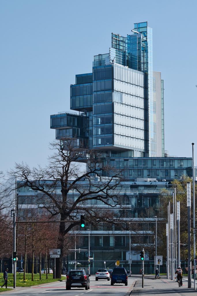 Ein modernes, hohes Gebäude mit einer auffälligen, abstrakten Fassade aus Glas und Stahl dominiert die Szene. Die Architektur ist durch viele unregelmäßige, schachbrettartig angeordnete Glaswürfel gekennzeichnet, die in verschiedenen Höhen und Winkeln aus dem Hauptkörper des Gebäudes herausragen. Einige Abschnitte sind mit vertikalen, silbrig-weißen Paneelen verkleidet, die mit den transparenten Glasflächen kontrastieren. Die obere Spitze des Gebäudes ist von spitz zulaufenden, turmartigen Glaselementen gekrönt, die in den klaren blauen Himmel ragen. Vor dem Gebäude erstreckt sich eine breite Straße mit mehrspurigem Verkehr. Mehrere Autos, darunter ein dunkler SUV und ein blauer Kleinwagen, fahren auf der Straße. Am rechten Fahrbahnrand sind Fußgänger und Radfahrer zu sehen, was auf ein belebtes städtisches Umfeld hindeutet. Links von der Straße befinden sich Bäume, von denen einige kahl sind und braune Äste aufweisen, was auf eine frühlingshafte Jahreszeit hindeutet.