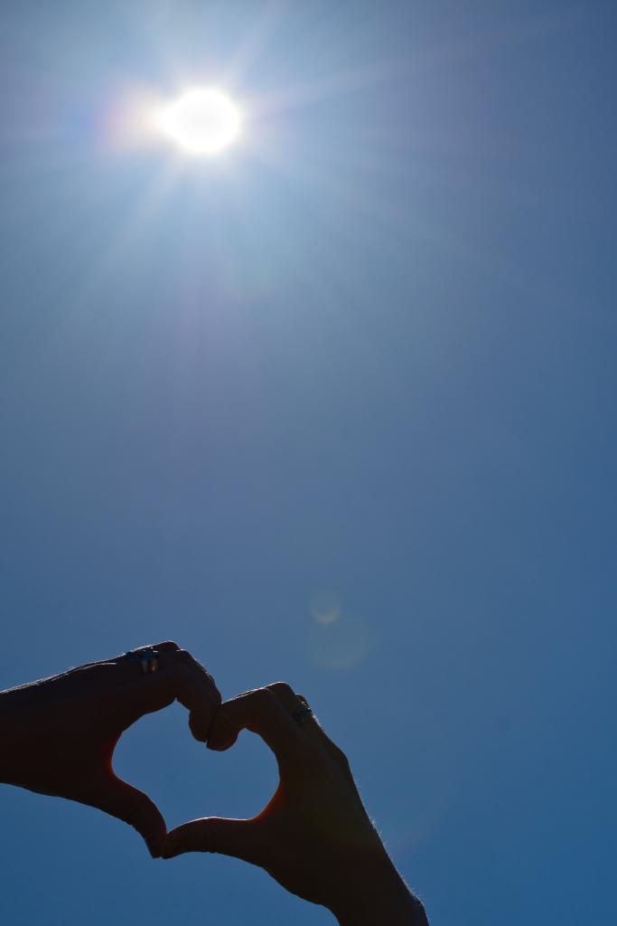 Silhouetten von zwei Händen, die geformt einen Herzsymbol vor einem hellblauen Himmel mit der Sonne bilden.