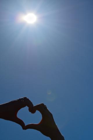 Silhouetten von zwei Händen, die geformt einen Herzsymbol vor einem hellblauen Himmel mit der Sonne bilden.