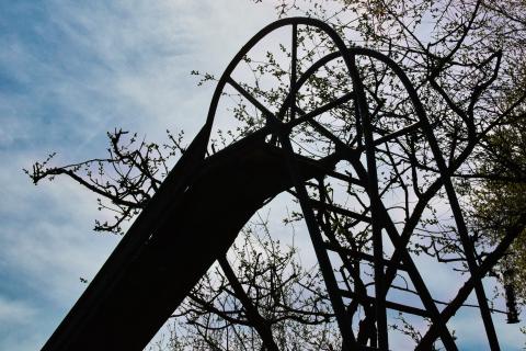 Eine dunkle Metallstruktur, die wie eine Rutsche aussieht, ragt gegen einen bewölkten Himmel auf. Sie ist von kahlen Ästen und einigen jungen Blättern umgeben, die von Bäumen stammen, die teilweise mit der Struktur verwachsen zu sein scheinen. Die Perspektive ist von unten aufgenommen, was der Struktur eine imposante Größe verleiht. Einzelne Elemente der Metallkonstruktion, wie gebogene Träger und Gitter, sind als Silhouetten gegen den hellen Himmel zu erkennen. Im Hintergrund sind deutliche Wolkenformationen zu sehen.