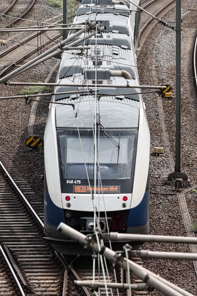 Eine moderne weiße und blaue Elektrolokomotive mit der Nummer 648 475 und dem Fahrtziel Hannover Hbf und der Linie RB38 fährt auf einem Gleis. Die Lok ist mit Oberleitungen verbunden, die sich über dem Zug und den Bahnschienen erstrecken. Die Bahnschienen sind von Schotter bedeckt und von Masten mit gelb-schwarzen Warnschildern gesäumt. Die Aufnahme wurde von oben gemacht und fokussiert auf die Front des Zuges.