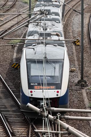 Eine moderne weiße und blaue Elektrolokomotive mit der Nummer 648 475 und dem Fahrtziel Hannover Hbf und der Linie RB38 fährt auf einem Gleis. Die Lok ist mit Oberleitungen verbunden, die sich über dem Zug und den Bahnschienen erstrecken. Die Bahnschienen sind von Schotter bedeckt und von Masten mit gelb-schwarzen Warnschildern gesäumt. Die Aufnahme wurde von oben gemacht und fokussiert auf die Front des Zuges.