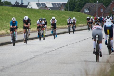 Eine Gruppe von Radfahrern (wirklich rein männlich wegen Iron Man) fährt auf einer Straße, die von einer grünen Böschung gesäumt ist. Die Radfahrer tragen aerodynamische Helme und Rennrad-Bekleidung. Sie sind auf Rennrädern unterwegs und befinden sich in einer Rennsituation, erkennbar an der Formation und der Konzentration der Athleten. Im Hintergrund sind ein grüner Deich direkt neben der Straße, Wohnhäuser mit Ziegeldächern und Solarpanels zu sehen, sowie weitere Rennteilnehmer in der Ferne. Der Himmel ist bewölkt.