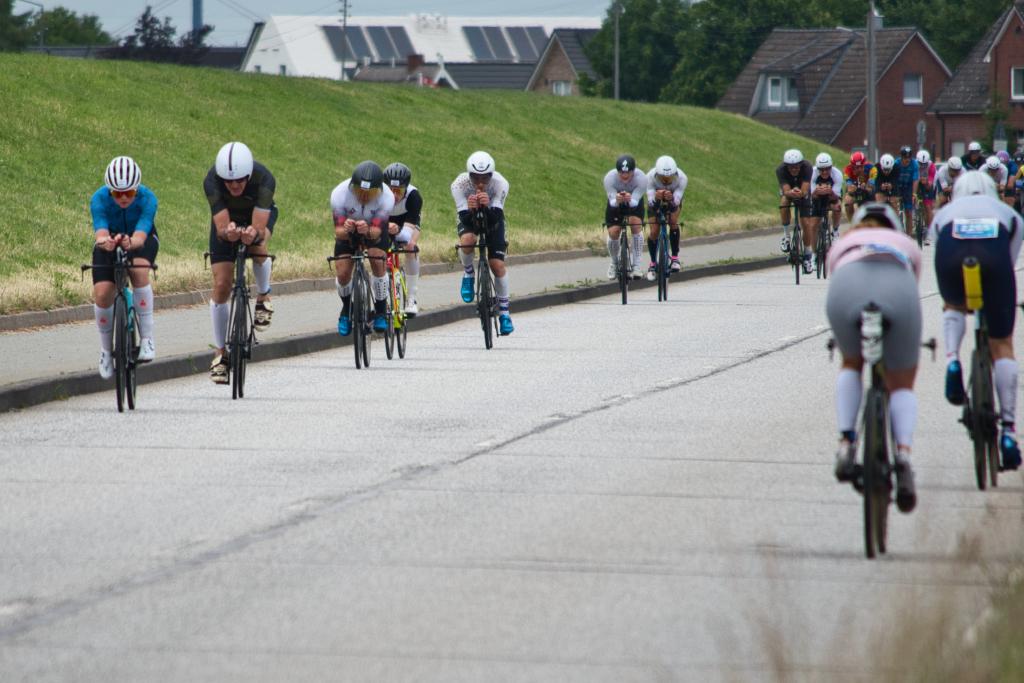 Eine Gruppe von Radfahrern (wirklich rein männlich wegen Iron Man) fährt auf einer Straße, die von einer grünen Böschung gesäumt ist. Die Radfahrer tragen aerodynamische Helme und Rennrad-Bekleidung. Sie sind auf Rennrädern unterwegs und befinden sich in einer Rennsituation, erkennbar an der Formation und der Konzentration der Athleten. Im Hintergrund sind ein grüner Deich direkt neben der Straße, Wohnhäuser mit Ziegeldächern und Solarpanels zu sehen, sowie weitere Rennteilnehmer in der Ferne. Der Himmel ist bewölkt.