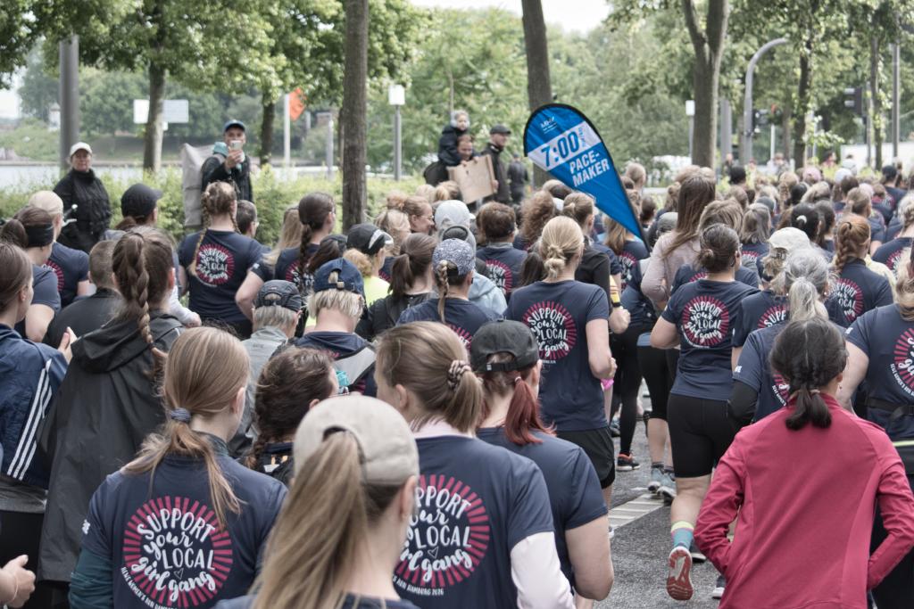 Eine große Gruppe von Frauen nimmt an einem Lauf teil, viele tragen dunkelblaue T-Shirts mit der Aufschrift "SUPPORT YOUR LOCAL GIRL GANG". Im Hintergrund ist eine blaue Fahne mit der Aufschrift "7:00 PACE MAKER" zu sehen. Die Teilnehmerinnen sind im Begriff, loszulaufen oder laufen bereits. Die Szene spielt sich im Freien ab, umgeben von Bäumen und gegen eine leicht unscharfe Kulisse von Gebäuden.