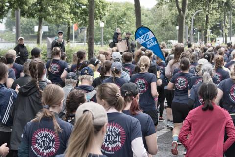 Eine große Gruppe von Frauen nimmt an einem Lauf teil, viele tragen dunkelblaue T-Shirts mit der Aufschrift "SUPPORT YOUR LOCAL GIRL GANG". Im Hintergrund ist eine blaue Fahne mit der Aufschrift "7:00 PACE MAKER" zu sehen. Die Teilnehmerinnen sind im Begriff, loszulaufen oder laufen bereits. Die Szene spielt sich im Freien ab, umgeben von Bäumen und gegen eine leicht unscharfe Kulisse von Gebäuden.