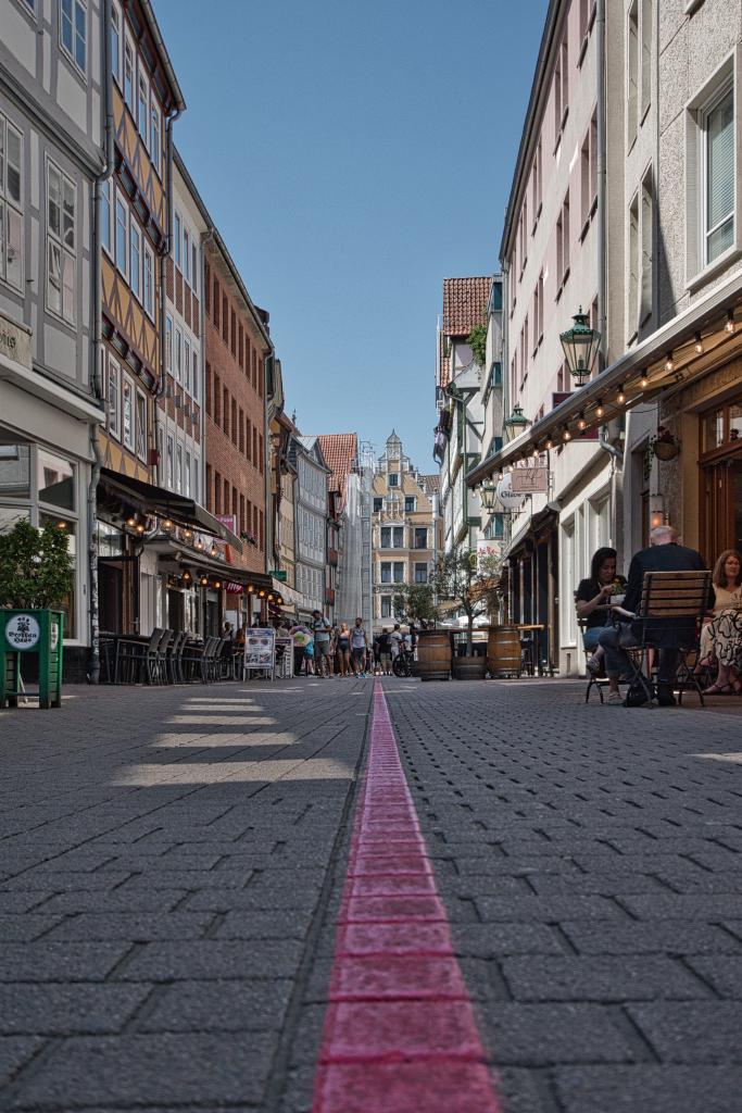 Die Kamera des Fotografen blickt von unten entlang einer gepflasterten Fußgängerzone direkt in die Mitte einer Häusergasse. Ein markanter, pinkfarbener Streifen verläuft mittig auf dem Boden geradeaus und führt den Blick des Betrachters in die Ferne. Links und rechts säumen historische Fachwerkhäuser und neuere Gebäude die Gasse, in denen sich zahlreiche Straßencafés und Restaurants befinden. Auf der rechten Seite sitzen Gäste entspannt an Tischen vor den Lokalen und genießen das sonnige Wetter bei blauem Himmel. Im Hintergrund endet die Sichtachse an einer imposanten Fassade eines älteren Gebäudes, während einige Passanten in der Ferne zu sehen sind.