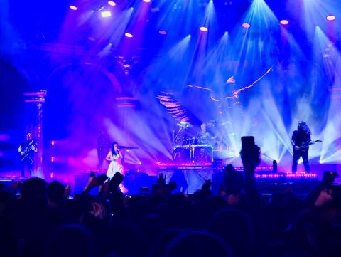 Bühnenaufnahme einer Band mit Sängerin, Gitarristen und Schlagzeuger vor Publikum und bunten Bühnenlichtern.