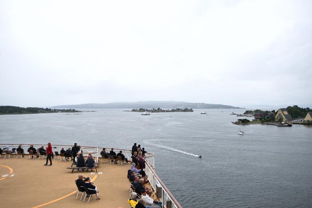 Menschen auf dem Deck eines Kreuzfahrtschiffes blicken in Norwegen unter einem bewölkten, grauen Himmel auf eine ruhige Bucht mit kleinen Booten, Inseln und Gebäuden.