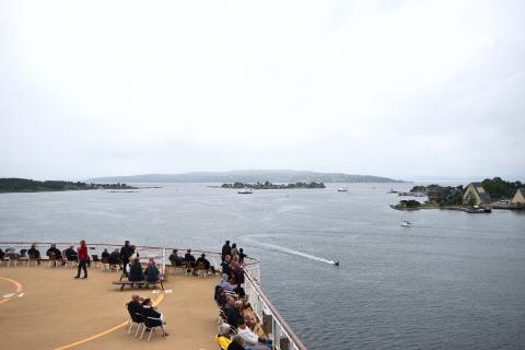 Menschen auf dem Deck eines Kreuzfahrtschiffes blicken in Norwegen unter einem bewölkten, grauen Himmel auf eine ruhige Bucht mit kleinen Booten, Inseln und Gebäuden.