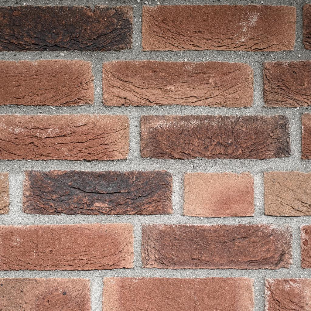 Nahaufnahme einer Klinkerwand mit rot- bis dunkelbraunen, rissig strukturierten Ziegeln in versetzten Reihen, verbunden durch hellgraue, körnige Mörtelfugen